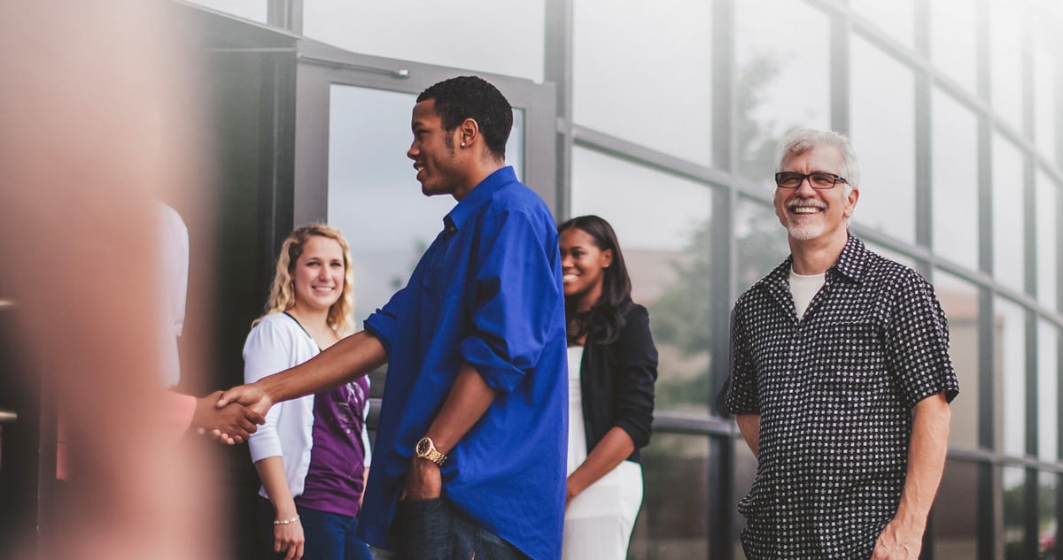 A group of diverse people smiles and shakes hands outside a modern building with large glass windows, suggesting a professional or social meeting.