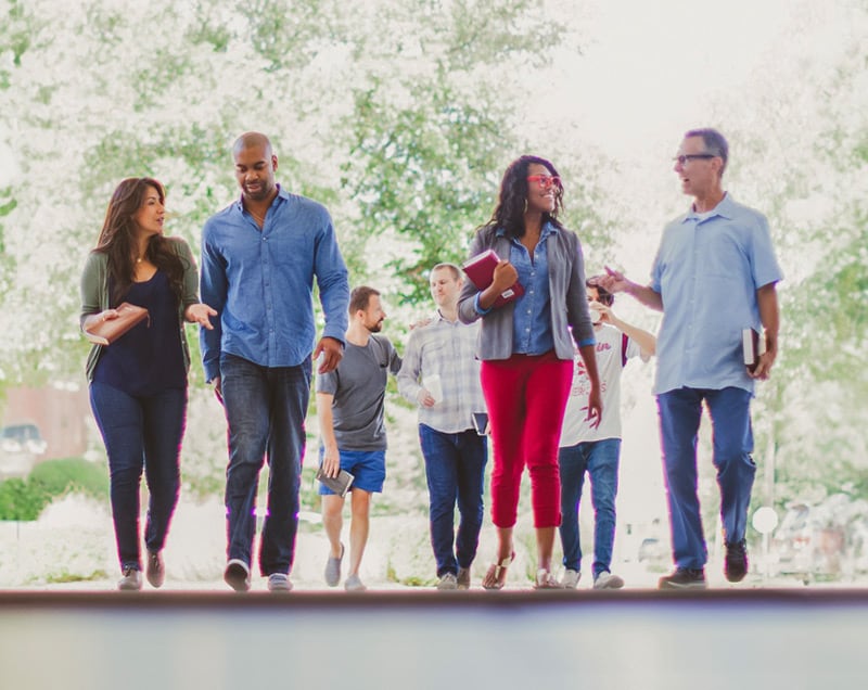 A diverse group of people walks and talks together outdoors, some holding books, in a casual professional setting.