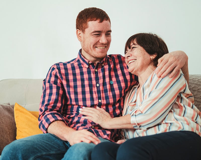 A smiling man and woman sit closely together on a couch, embracing in a warm, familial moment.