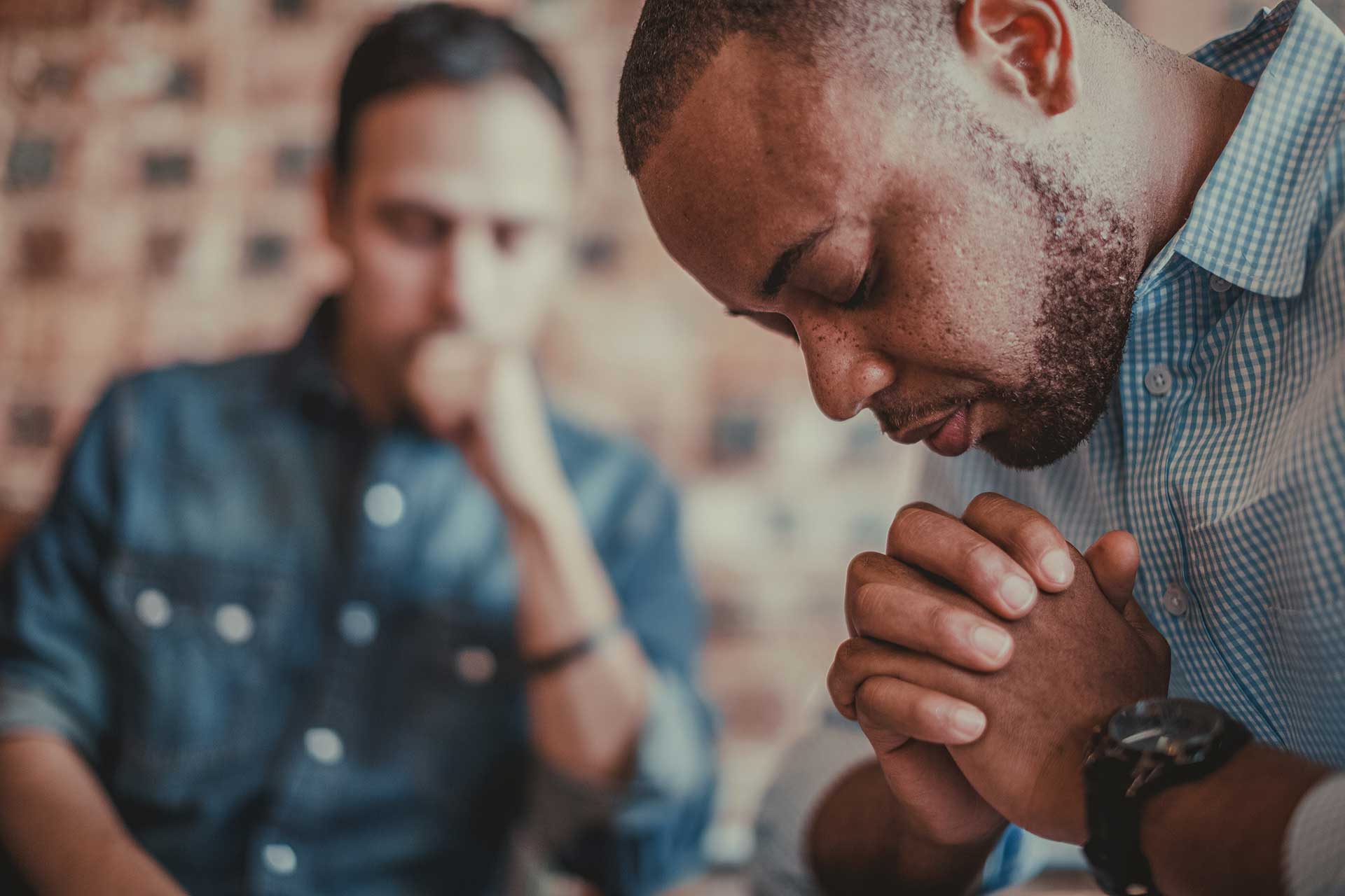 Two men sitting at a counselling session, one in front with hands clasped as if praying, the other blurred in the background