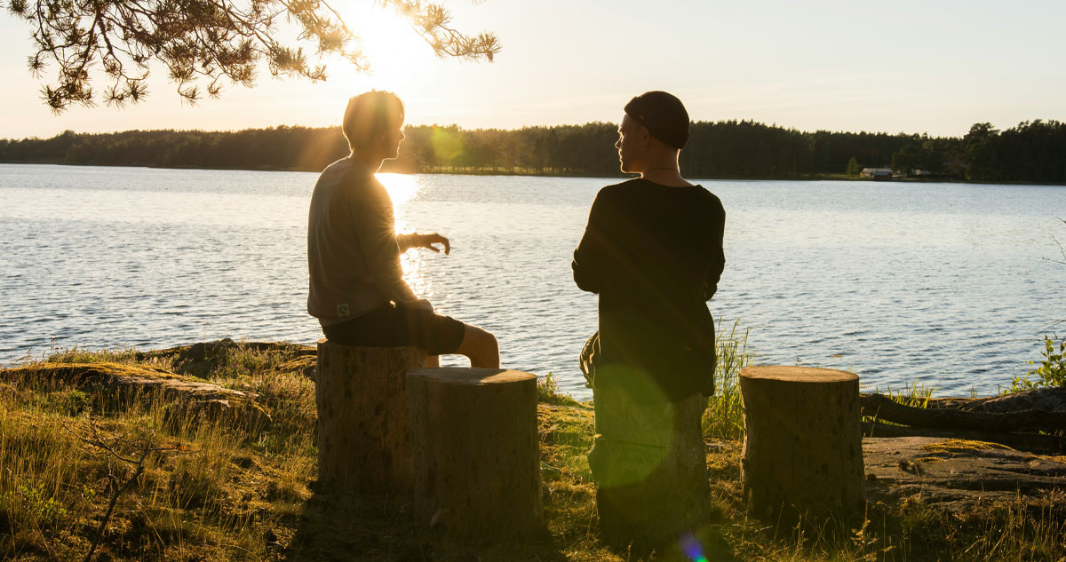 0179ADF6-83AF-47EF-A7BF-1CA41B0A076D_web Two people sitting on tree stumps, in kind conversation, with sun rising over lake in front.
